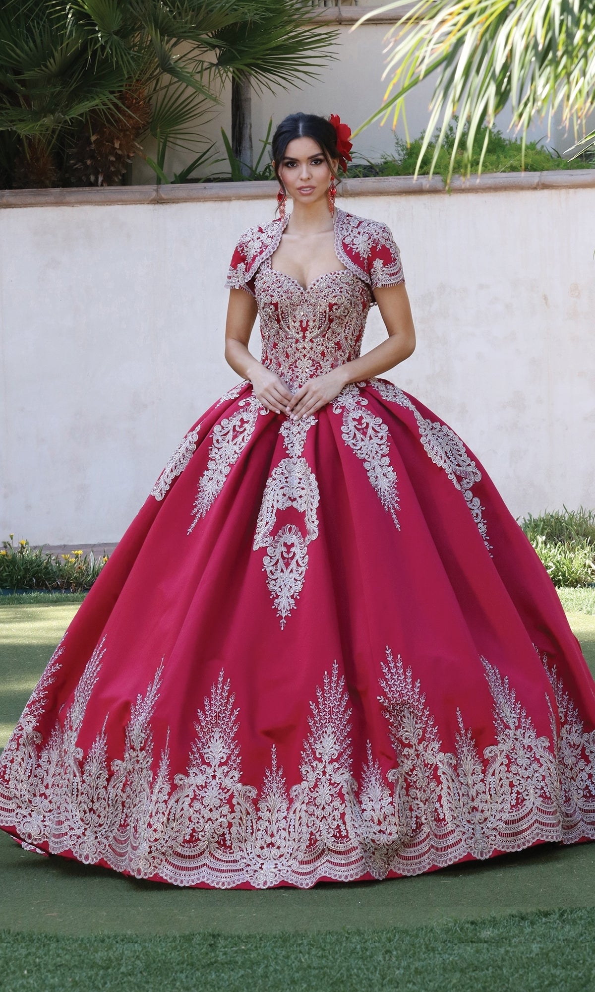A woman stands outdoors in the Quinceanera Dress 1494 by Dancing Queen, a bright red a-line ball gown with gold lace embroidery, hands clasped at her waist and a matching bolero finishing the look.